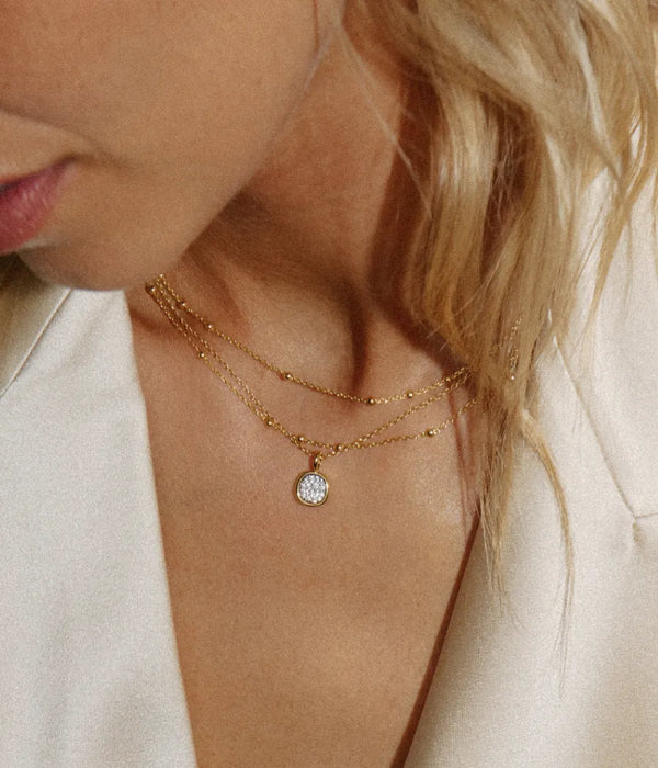 Close-up of a woman wearing a delicate gold double-layer necklace with a small oval clear gemstone pendant, paired with a cream satin blazer.